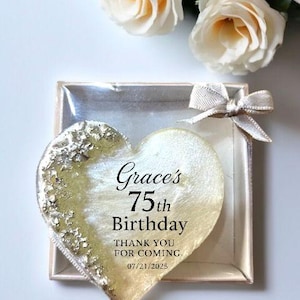 May include: A heart-shaped party favor with gold leaf accents and the text "Grace's 75th Birthday." The favor is in a clear box with a silver bow, and white roses are in the background.