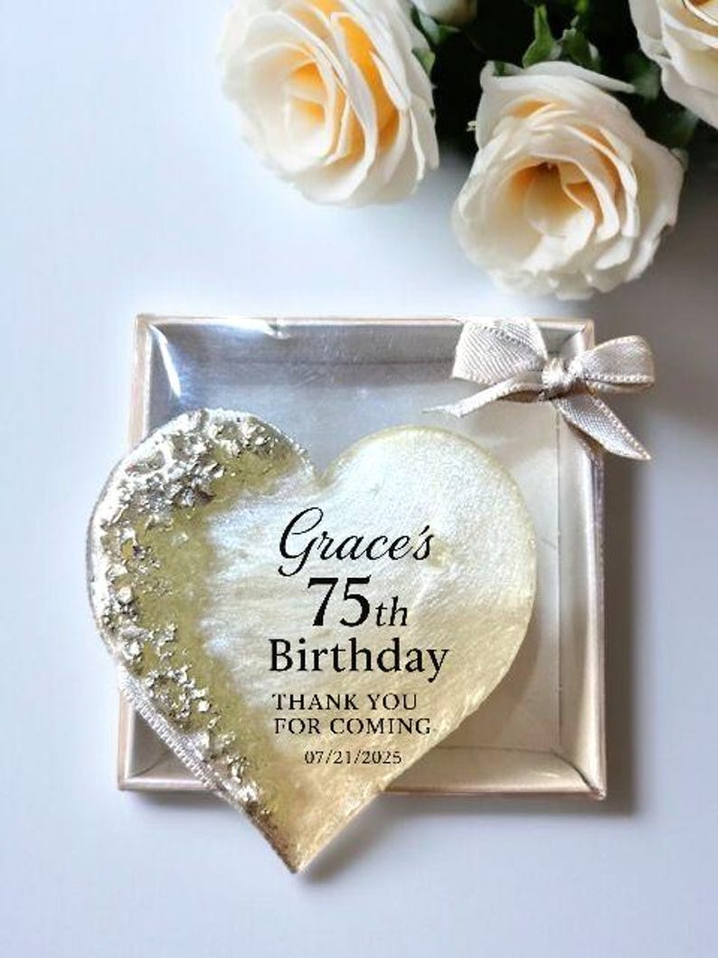 May include: A heart-shaped party favor with gold leaf accents and the text "Grace's 75th Birthday." The favor is in a clear box with a silver bow, and white roses are in the background.