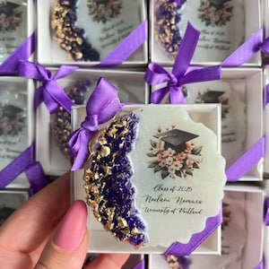 Pode incluir: Lembrancinhas de formatura em caixas brancas amarradas com fitas roxas. Cada caixa cont&eacute;m um porta-copos com um design roxo e dourado, um gr&aacute;fico de chap&eacute;u de formatura e o texto "Class of 2025".