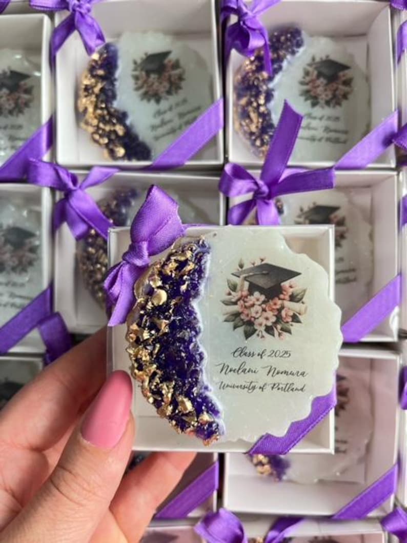 Pode incluir: Lembrancinhas de formatura em caixas brancas amarradas com fitas roxas. Cada caixa cont&eacute;m um porta-copos com um design roxo e dourado, um gr&aacute;fico de chap&eacute;u de formatura e o texto "Class of 2025".