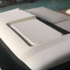 May include: A close-up of a white, rectangular foam component with a recessed, rectangular section. The component appears to be part of a larger structure, possibly automotive or furniture related. The surface is smooth and clean.