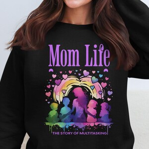 Puede incluir: Sudadera negra con un gráfico morado de "Mom Life". El gráfico presenta una ilustración al estilo acuarela de una madre y sus hijos con corazones y el texto "The Story of Multitasking".