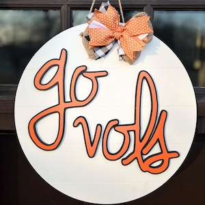 May include: A white round wooden door hanger with the words "go vols" in orange and black lettering. A large orange and white polka dot bow with a burlap bow is attached to the top.