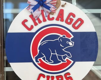 May include: A white, blue, and red round wooden door hanger with the Chicago Cubs logo. The logo features a red and blue 'C' with a blue bear inside. The words 'Chicago' and 'Cubs' are in red letters.
