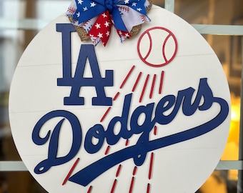 May include: A white round wooden door hanger with a blue and red baseball themed design. The design features the letters "LA" and the word "Dodgers" in blue with red rays of light behind them. A red baseball is in the upper right corner of the design. A red, white, and blue bow with stars is at the top of the door hanger.