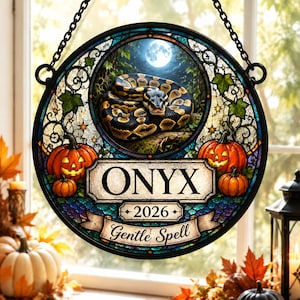 May include: A circular stained glass suncatcher featuring a coiled snake under a full moon, flanked by jack-o'-lanterns. The center displays the text ONYX, 2026, and Gentle Spell on a banner. It hangs from a black metal chain.
