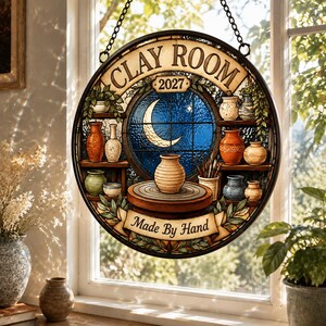 May include: A circular stained glass suncatcher featuring the text CLAY ROOM 2027 and MADE BY HAND. The design depicts a pottery wheel with a clay pot, surrounded by shelves of various ceramic vases and a blue window with a crescent moon.