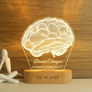 Op de afbeelding: Een oplichtende hersenvormige nachtlamp met de naam "Brand Elmegen" en de titel "NEUROPSYCHOLOGIST" gegraveerd. De lamp staat op een houten voet met de datum "02.16.2025". Het licht heeft een warme gele kleur.
