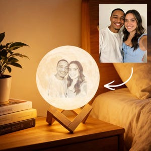 May include: A personalized moon lamp with a photo of a couple. The lamp is round and emits a soft, warm light. It sits on a wooden stand. A small photo of the couple is also visible in the upper right corner.