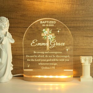 Puede incluir: Una luz de noche de bautizo personalizada con el nombre "Emma Grace" y la fecha "06-18-2025". El panel acrílico transparente presenta una cruz con detalles florales y un versículo bíblico, sobre una base de madera con luces LED.