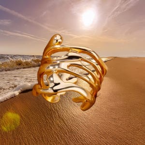 Peut inclure: Une bague dorée au design abstrait et fluide. La bague présente une forme organique avec plusieurs espaces ouverts. La scène de fond montre une plage avec la mer et un coucher de soleil.