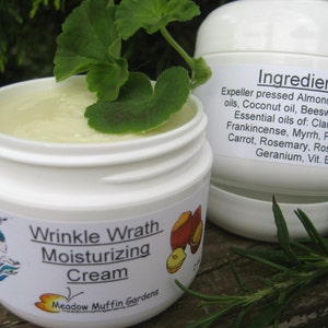 May include: A white jar with a green leaf on top. The jar is labeled "Wrinkle Wrath Moisturizing Cream" and "Meadow Muffin Gardens". The ingredients are listed on the side of the jar: "Expeller pressed Almond & Hazelnut oils, Coconut oil, Beeswax, Water, Essential oils of: Clary sage, Frankincense, Myrrh, Patchouli, Carrot, Rosemary, Rosewood, Geranium, Vit. E"
