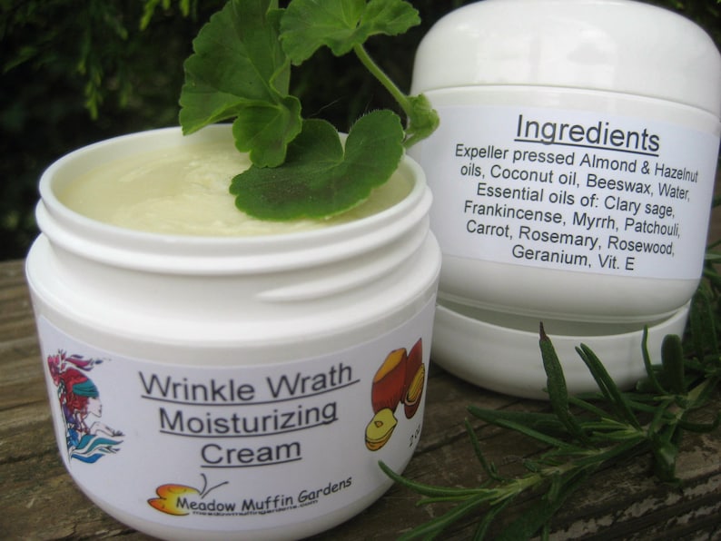 May include: A white jar with a green leaf on top. The jar is labeled "Wrinkle Wrath Moisturizing Cream" and "Meadow Muffin Gardens". The ingredients are listed on the side of the jar: "Expeller pressed Almond & Hazelnut oils, Coconut oil, Beeswax, Water, Essential oils of: Clary sage, Frankincense, Myrrh, Patchouli, Carrot, Rosemary, Rosewood, Geranium, Vit. E"