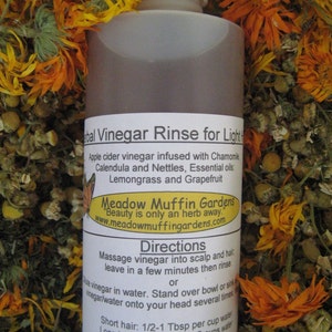 Może przedstawiać: Brązowa szklana butelka z białym korkiem, z etykietą "Herbal Vinegar Rinse for Light Hair". Etykieta zawiera listę składnik&oacute;w, w tym ocet jabłkowy, rumianek, nagietek, pokrzywę, trawy cytrynowej i grejpfruta. Butelka jest otoczona suszonymi kwiatami.