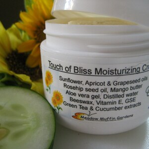 May include: A white jar with a white lid labeled "Touch of Bliss Moisturizing Cream". The label lists ingredients including sunflower, apricot, grapeseed oils, rosehip seed oil, mango butter, aloe vera gel, distilled water, beeswax, vitamin E, GSE, green tea and cucumber extracts. The label also includes the text "Meadow Muffin Gardens".