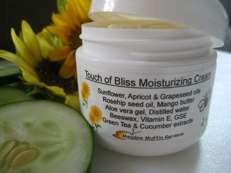 May include: A white jar with a white lid labeled "Touch of Bliss Moisturizing Cream". The label lists ingredients including sunflower, apricot, grapeseed oils, rosehip seed oil, mango butter, aloe vera gel, distilled water, beeswax, vitamin E, GSE, green tea and cucumber extracts. The label also includes the text "Meadow Muffin Gardens".