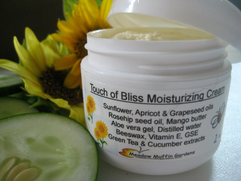 May include: White jar of Touch of Bliss Moisturizing Cream with a sunflower and green tea and cucumber extracts listed on the label. The jar is open and the cream is visible inside.