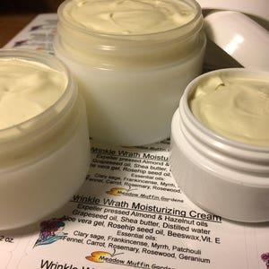 May include: Three glass jars filled with a white cream labeled "Wrinkle Wrath Moisturizing Cream". The label lists the ingredients, including almond oil, shea butter, and rosehip seed oil. The jars are on a surface with a printed label that says "Wrinkle Wrath Moisturizing Cream" and lists the ingredients.