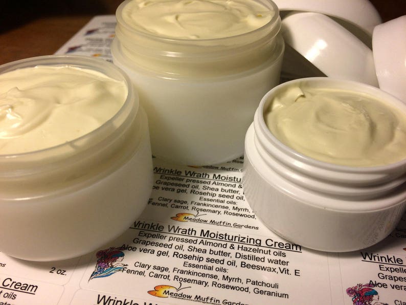 May include: Three glass jars filled with a white cream labeled "Wrinkle Wrath Moisturizing Cream". The label lists the ingredients, including almond oil, shea butter, and rosehip seed oil. The jars are on a surface with a printed label that says "Wrinkle Wrath Moisturizing Cream" and lists the ingredients.