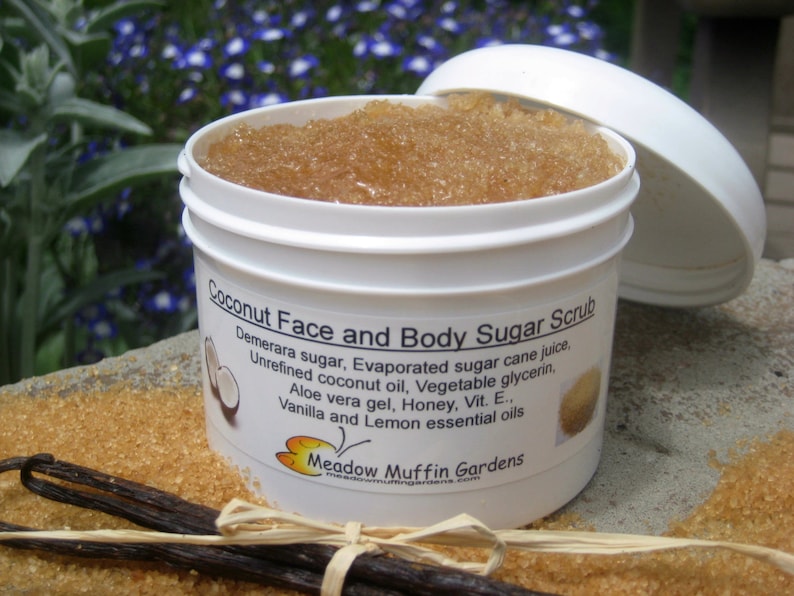 May include: A white jar with a brown sugar scrub inside. The label reads "Coconut Face and Body Sugar Scrub" and lists the ingredients: Demerara sugar, evaporated sugar cane juice, unrefined coconut oil, vegetable glycerin, aloe vera gel, honey, vitamin E, vanilla and lemon essential oils. The jar is sitting on a surface covered in brown sugar crystals.
