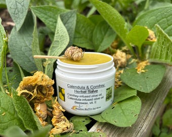 May include: A white jar of Calendula & Comfrey Herbal Salve with a yellow lid. The jar is sitting on a bed of green leaves and yellow flowers. The label on the jar reads "Calendula & Comfrey Herbal Salve", "Comfrey-infused olive oil, Calendula-infused olive oil", "Beeswax, vit. E", and "Meadow Muffin Gardens".