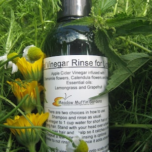 Może przedstawiać: Zielona butelka ze szkła z czarną zakrętką i białą etykietą z napisem "Vinegar Rinse for Light Hair". Etykieta zawiera r&oacute;wnież tekst "Ocet jabłkowy z dodatkiem kwiat&oacute;w rumianku, kwiat&oacute;w nagietka i cytryny. Olejki eteryczne: trawa cytrynowa i grejpfrut". Butelka stoi na zielonej trawie i ż&oacute;łtych kwiatach.