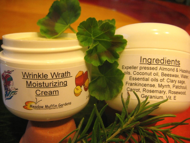 May include: Two white jars of wrinkle wrath moisturizing cream. The jar on the left has a colorful illustration of a woman and fruit. The jar on the right has a list of ingredients including almond, coconut, beeswax, clary sage, frankincense, myrrh, patchouli, carrot, rosemary, rosewood, geranium, and vitamin E.