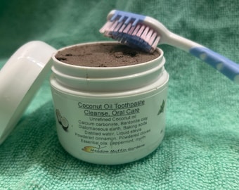 May include: A white plastic jar of dark brown coconut oil toothpaste with a detached white lid and a blue and white toothbrush resting on top. The label reads Coconut Oil Toothpaste Cleanse, Oral Care, listing ingredients and Meadow Muffin Gardens.