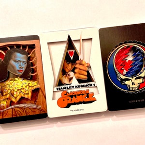 May include: Three framed images with a 3D effect. The first image features a woman with dark hair and a yellow and gold patterned top. The second image features a scene from the movie "A Clockwork Orange" with the text "Stanley Kubrick's A Clockwork Orange". The third image features a red, white, and blue skull with the text "© 2016 & TM GOP".