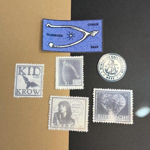 May include: A collection of stickers and stamps with a vintage aesthetic. Includes a blue patch with a wishbone and the words "WISHBONE CONAN GRAY". Other items feature a bird, a portrait, and the words "KID KROW", "WISH BONE", "FOUND HEAVEN", and "SUPERACHE".