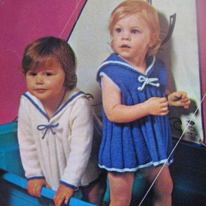 Könnte beinhalten: Zwei Kinder in Matrosen-gestrickten Outfits stehen in einem blauen Plastikboot. Das Mädchen links trägt einen weißen Pullover mit blauen Verzierungen und einer blauen Schleife. Das Mädchen rechts trägt ein blaues Kleid mit weißen Verzierungen und einer weißen Schleife.