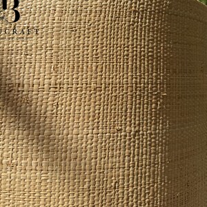 May include: Close-up of a cylindrical lampshade with a woven, textured design. The lampshade is a light tan color and has a repeating pattern. The brand name "BOUCRAFT" is visible on the side. The background shows a blue sky and tree branches.