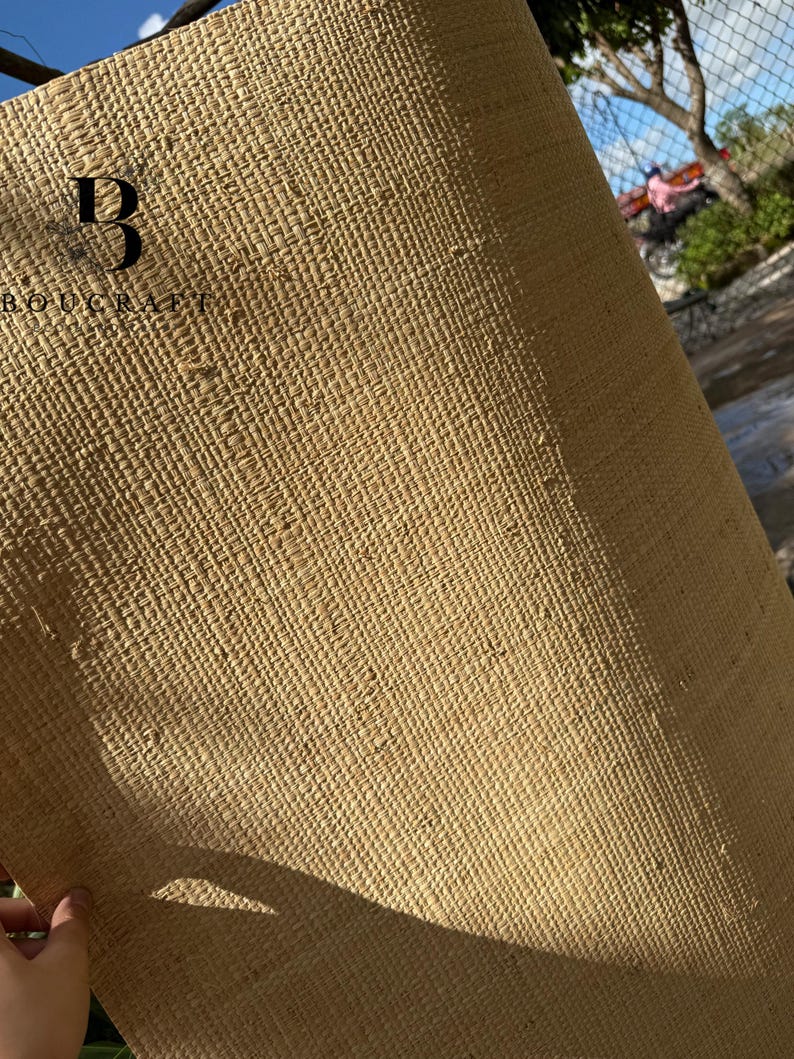 May include: A roll of woven natural fiber material in a light tan color. The material has a textured, grid-like pattern. The upper left corner displays the text "BOUGCRAFT" with a stylized "B" logo. The material is being held by a hand.
