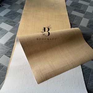May include: A rolled-up, rectangular, natural-colored jute runner rug with a white backing. The rug is partially unrolled, revealing the textured weave of the jute and the white, dotted backing. The logo "BOUCRAFT" is visible.