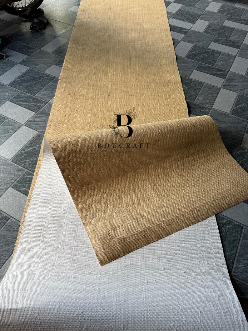 May include: A rolled-up, rectangular, natural-colored jute runner rug with a white backing. The rug is partially unrolled, revealing the textured weave of the jute and the white, dotted backing. The logo "BOUCRAFT" is visible.
