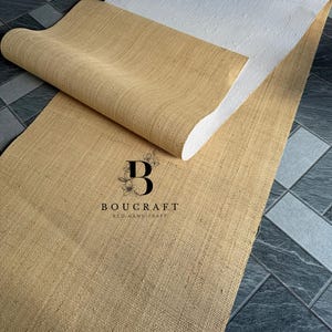 May include: A rolled-up, tan-colored jute rug with a white backing is displayed on a tiled floor. The rug is partially unrolled, revealing its textured surface. The logo "BOUCRAFT ECO HANDICRAFT" is visible on the rug.