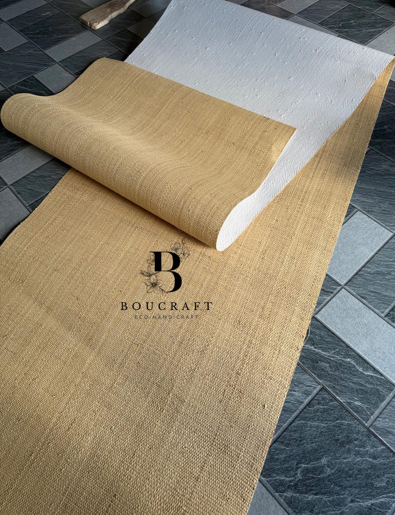 May include: A rolled-up, tan-colored jute rug with a white backing is displayed on a tiled floor. The rug is partially unrolled, revealing its textured surface. The logo "BOUCRAFT ECO HANDICRAFT" is visible on the rug.