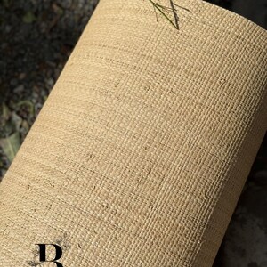 May include: A rolled-up, beige woven material with a textured surface. The material appears to be made of natural fibers. The logo "BOUCRAFT ECO HANDICRAFT" is visible at the bottom. A few green stems with red flowers are visible.
