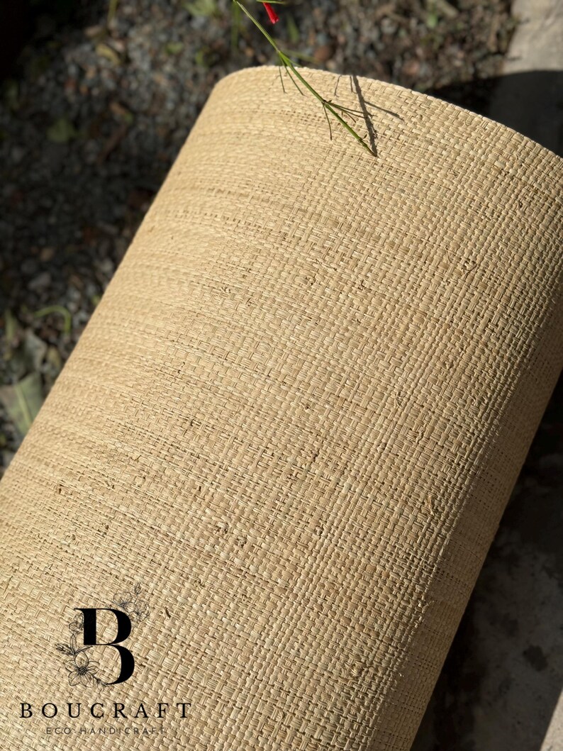 May include: A rolled-up, beige woven material with a textured surface. The material appears to be made of natural fibers. The logo "BOUCRAFT ECO HANDICRAFT" is visible at the bottom. A few green stems with red flowers are visible.