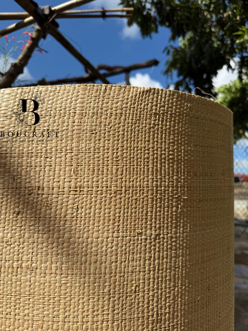May include: Close-up of a cylindrical lampshade with a woven, textured design. The lampshade is a light tan color and has a repeating pattern. The brand name "BOUCRAFT" is visible on the side. The background shows a blue sky and tree branches.