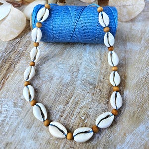 May include: A necklace made of white seashells and small wooden beads strung on blue thread. The necklace is displayed on a wooden surface next to a spool of blue thread and decorative elements.