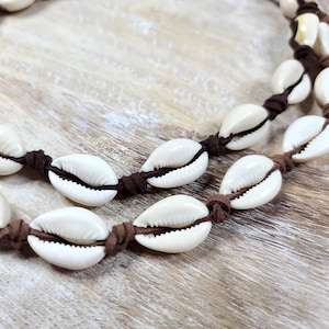 Puede incluir: Dos collares hechos de conchas de cauri blancas ensartadas en un cordón de gamuza marrón. Las conchas tienen forma ovalada con un acabado natural y brillante. El cordón está anudado entre cada concha.
