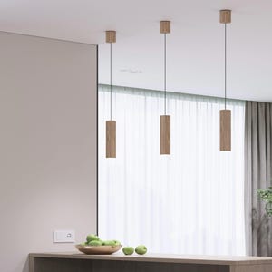 Könnte beinhalten: Drei moderne Pendelleuchten mit zylindrischen Holzschirmen hängen von der Decke. Die Leuchten sind an dünnen schwarzen Schnüren aufgehängt. Der Raum hat eine neutrale Farbpalette mit einer hellen Wand und einer Holztheke. Auf der Theke steht eine Schale mit grünen Äpfeln.