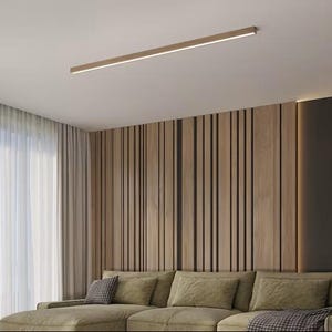 May include: A contemporary living room with a long, rectangular recessed light fitting on the ceiling. The room features a light-coloured sofa, wooden panelling, and sheer curtains. The colour scheme is neutral, with shades of beige and brown.