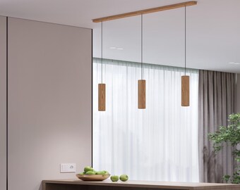 May include: Three wooden pendant lights hanging from a wooden beam. The cylindrical lights are suspended by black cords. The lights are in a modern kitchen setting with a neutral color palette.