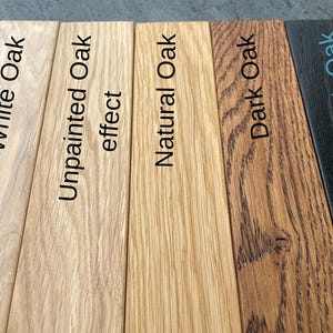 May include: A close-up of five wooden planks, each showcasing a different oak finish. The finishes include White Oak, Unpainted Oak effect, Natural Oak, Dark Oak, and Black Oak, with the names of each finish printed on the planks.