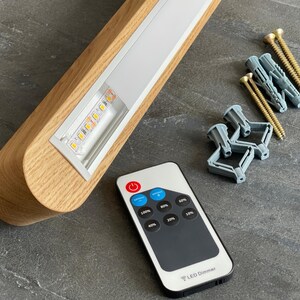 Light oak linear LED pendant light with visible LED strip, remote control, mounting hardware, and wall plugs. Complete set for easy installation and dimmable lighting control.