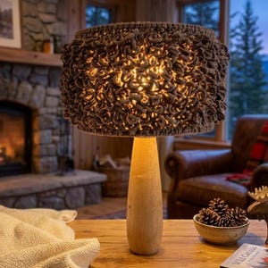 Könnte beinhalten: Eine dekorative Tischlampe mit hellbraunem Holzfuß und einem strukturierten, dunkelbraunen Lampenschirm. Die Lampe steht auf einem Holztisch mit einer Schale mit Tannenzapfen und einer Holzfigur eines Elchs. Ein Kamin und ein Sessel befinden sich im Hintergrund.