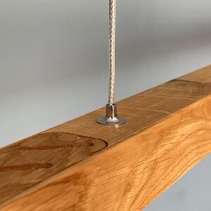Close-up of natural oak linear pendant light with stainless steel suspension cable and visible wood grain. Handmade modern LED chandelier in Scandinavian Japandi style.