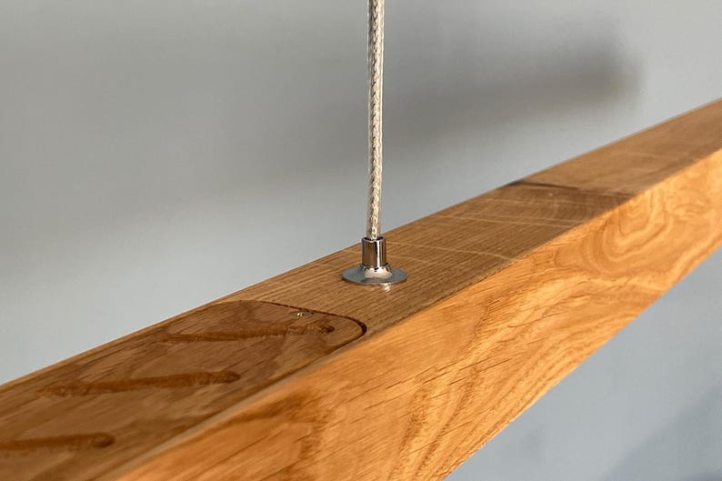 Close-up of natural oak linear pendant light with stainless steel suspension cable and visible wood grain. Handmade modern LED chandelier in Scandinavian Japandi style.